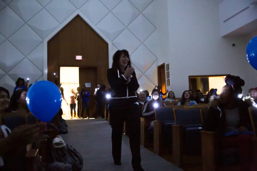 Image of President Mantella's entrance into program. Students use phone flashlights to light the aisle as she walks in to dark room